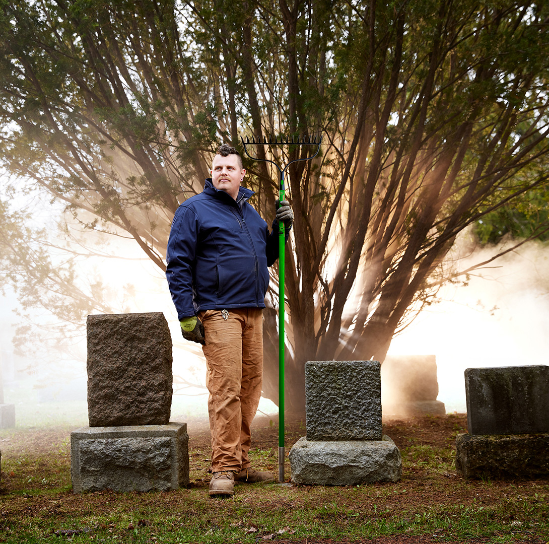 Jardinier du cimetière debout devant un arbre rétro-éclairé entouré de pierres tombales.